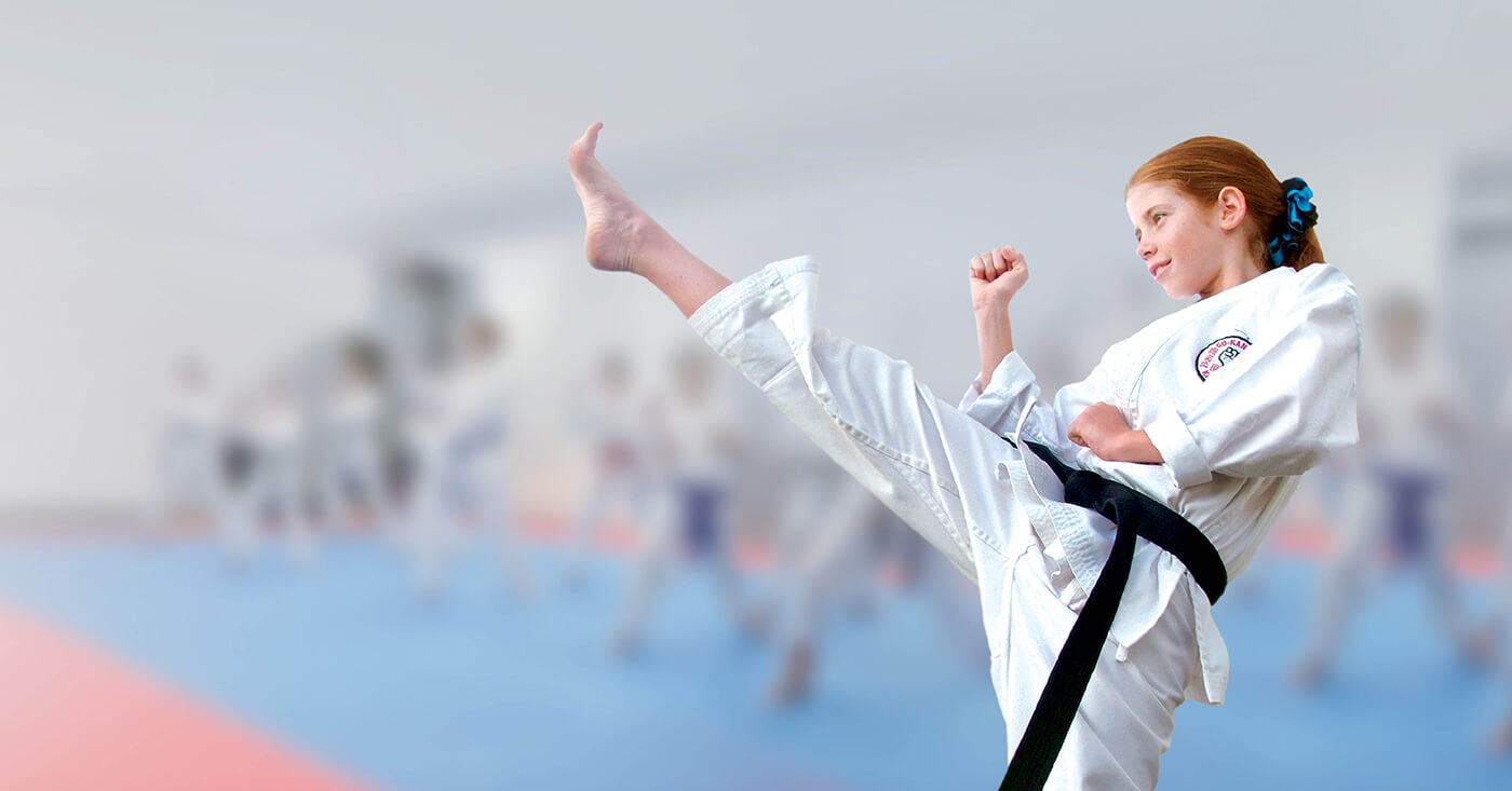 Girl in a gi and a black belt pictured side on kicking her leg into the air