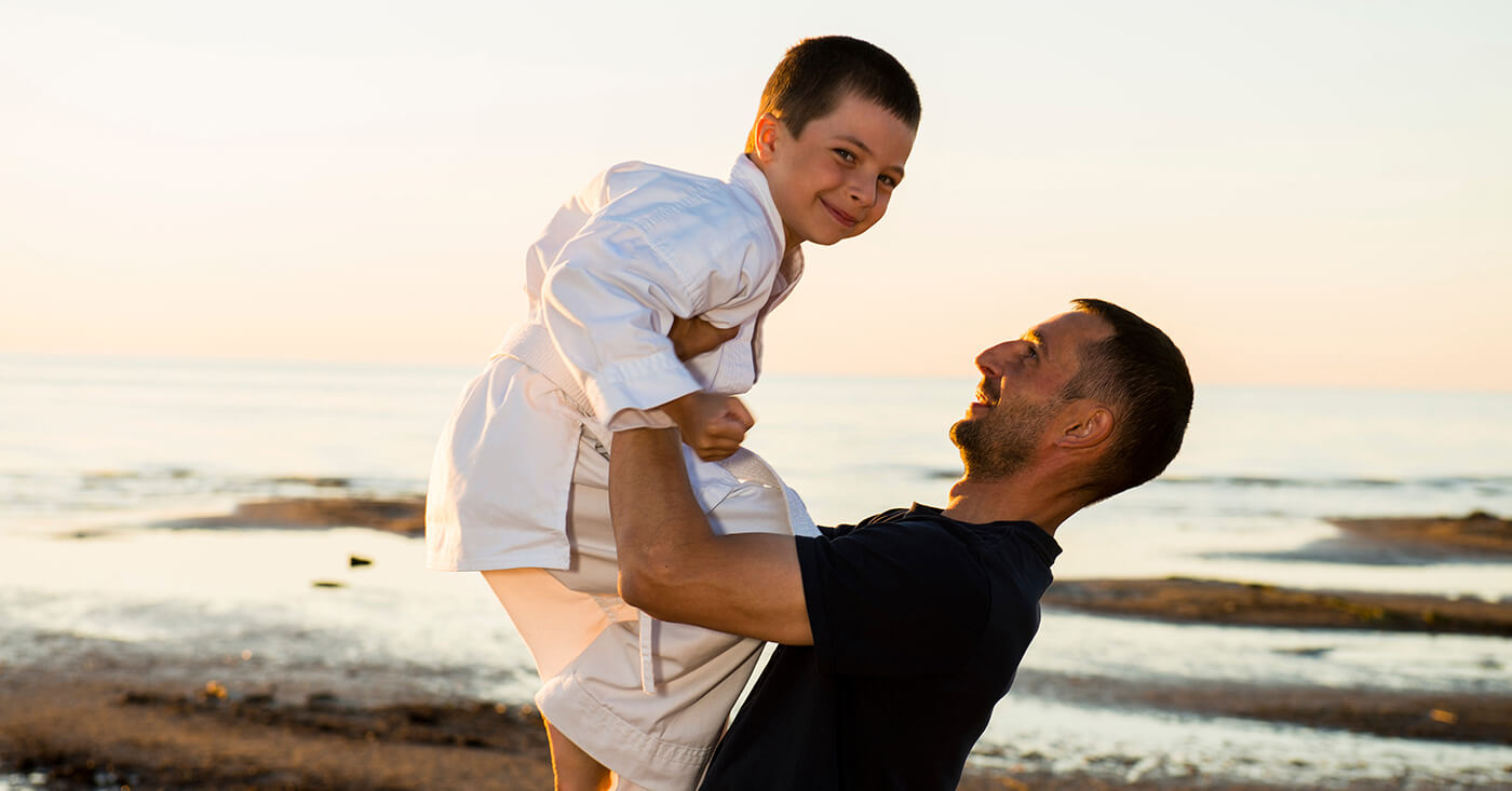 Father holding his son up in the air at the beach. The son is wearing a gi. kids doing karate